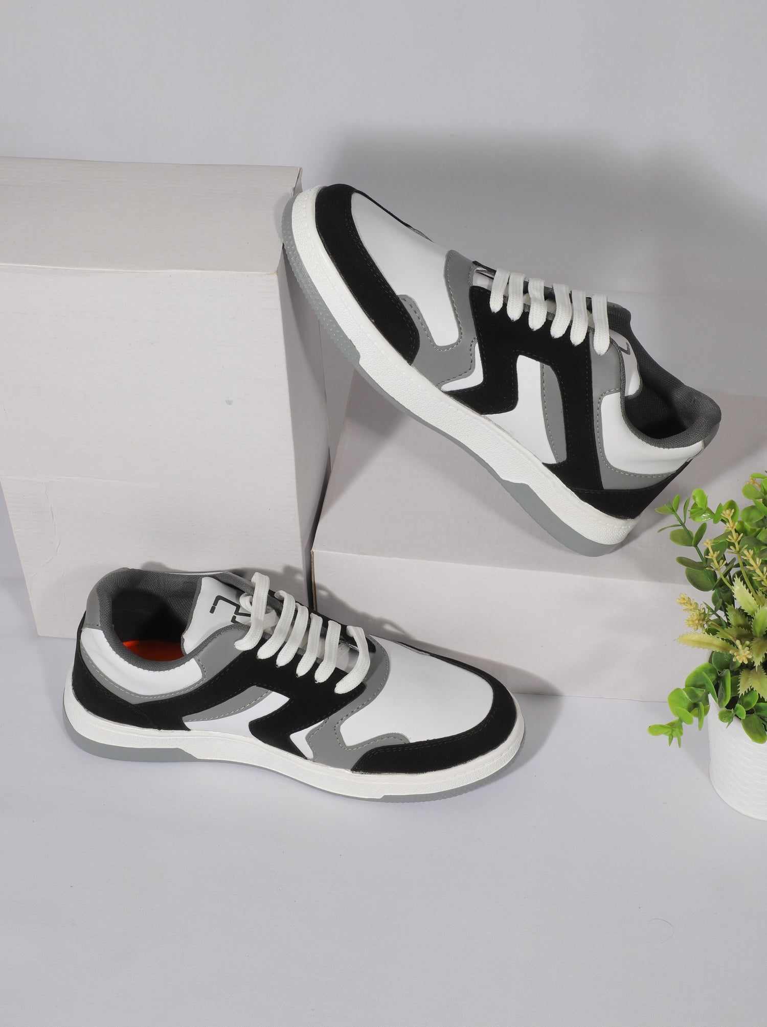 A pair of stylish black, white, and grey sneakers with white laces displayed on white boxes. The shoes have a modern, sporty design with contrasting panels and a clean, urban aesthetic
