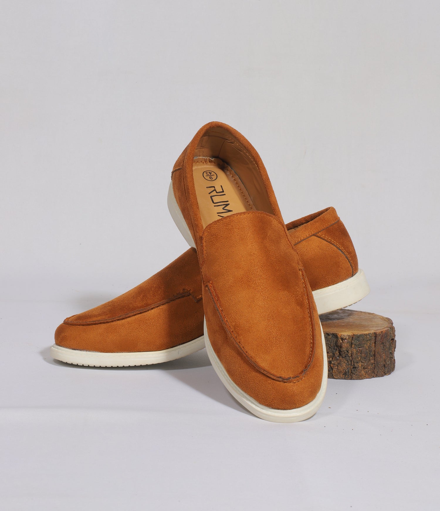 A pair of tan suede loafers with white soles, branded 'RUMANI', elegantly displayed on a white surface with a wooden stump accent. The loafers feature a minimalist design and soft finish, suited for casual or semi-formal wear.